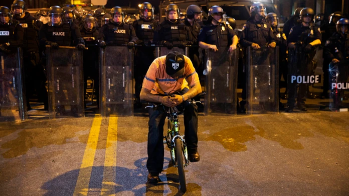 Muž sedí na bicykli pred policajnou líniou počas zákazu vychádzania v Baltimore.