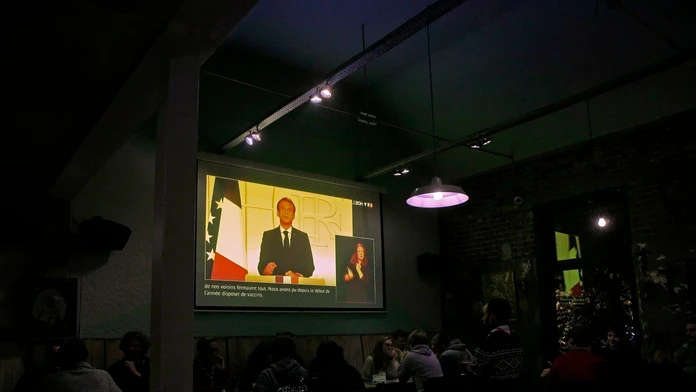 Ľudia sa zabávajú v bare a sledujú televíziu, kde francúzsky prezident Emmanuel Macron vystupuje s televíznym príhovorom.
