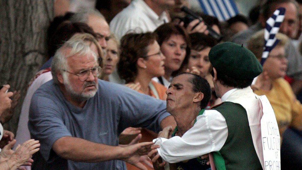 Neznámy muž v kostýme napadá favorita pretekov, brazílskeho atléta Vanderleia De Limu, na maratónskej trati v Aténach.